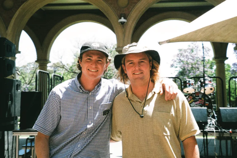 Tom Redden and Jack Stokes from Daybed Records smiling together in hats outdoors under archways at an Adelaide music venue or outdoor festival.